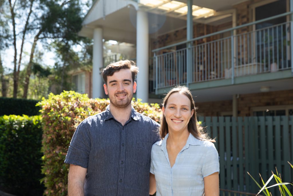 Laura and Finn standing in the front yard outside their apartment smiling into the camera