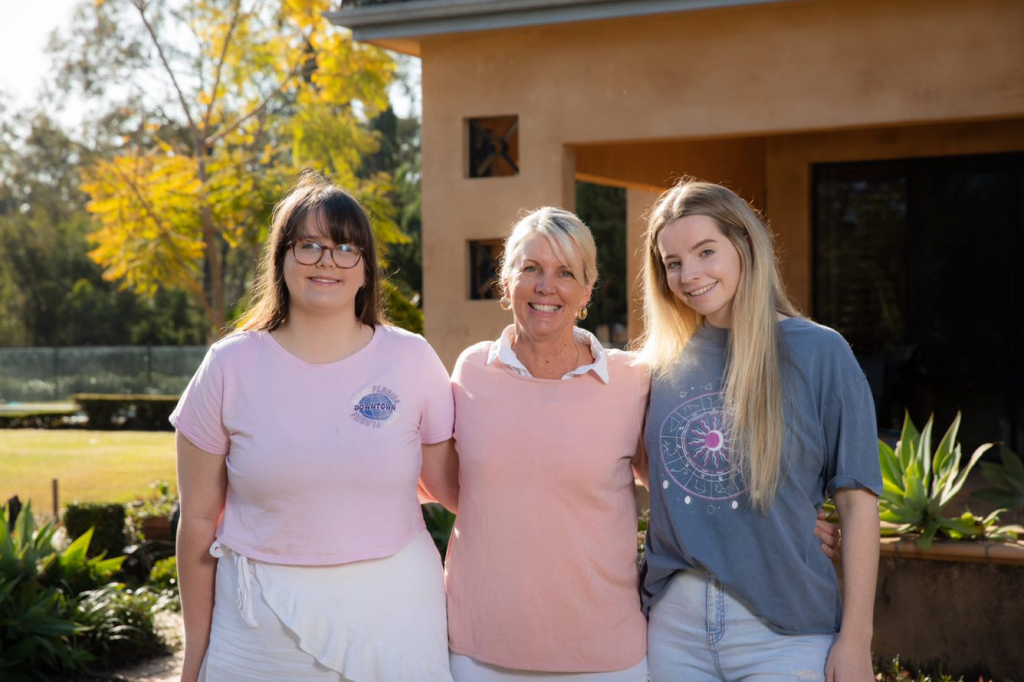 Heidi Morris and two of her daughters standing in their front yard smiling into the camera.