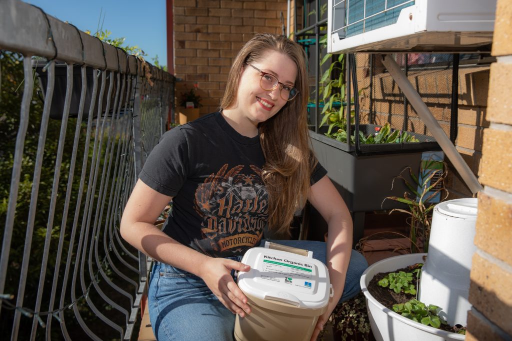 Young woman pours food waste from bucket into small earth worm compost farm on her balcony.