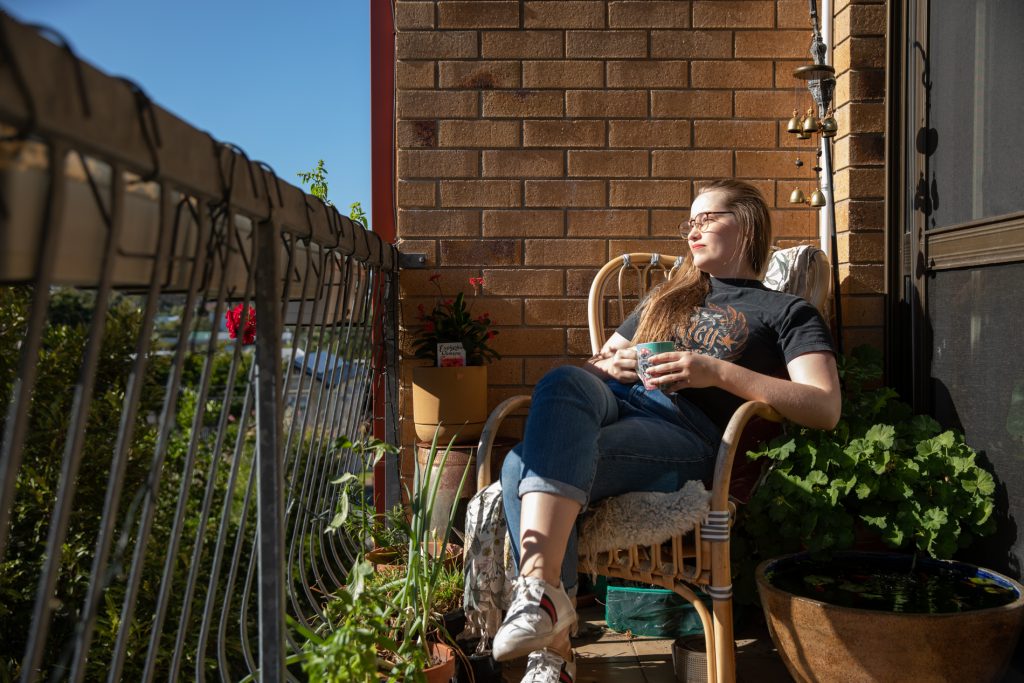 Young woman sits on a balcony with lots of greenery in the sun, looking outwards while holding a tea cup.