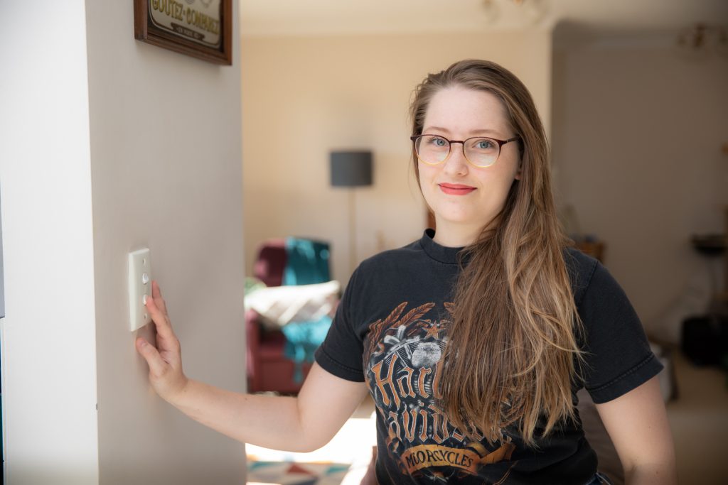 Young woman with glasses smiling into camera as she turns off lightswitch in her apartment.