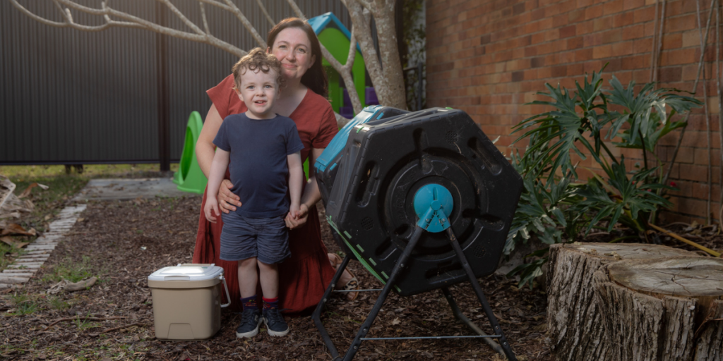 Emma Boyd kneeling on the ground, holding her toddler son who is standing up, next to the compost tumbler and smiling into the camera.