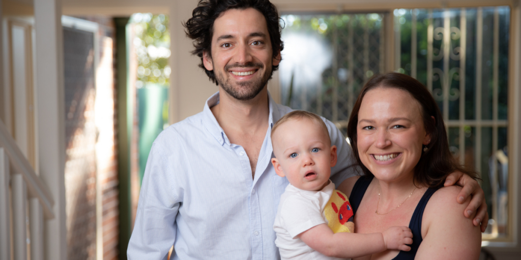 Cavdarski family standing in their living room, holding their baby son and smiling into the camera.