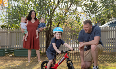 The Boyd family standing in their yard with Emma holding both twin baby boys and Brendan watching their other young son ride a bike.