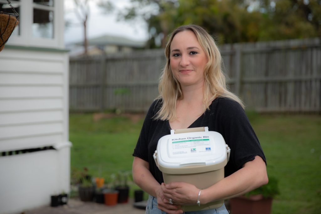 Karina standing in her yard, smiling into the camera while holding a compost caddy.