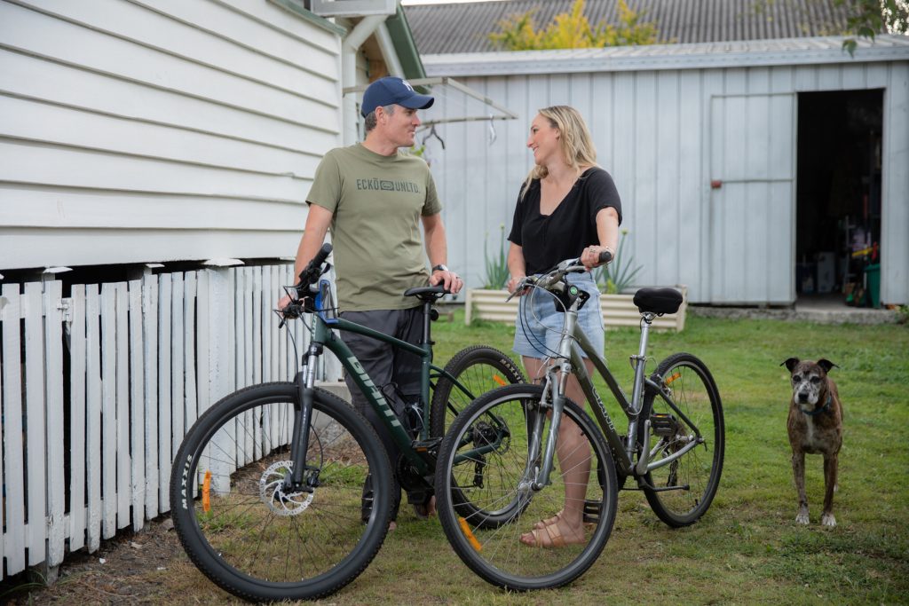 Brad and Karina walking their bikes in their front yard while smiling, looking at each other.