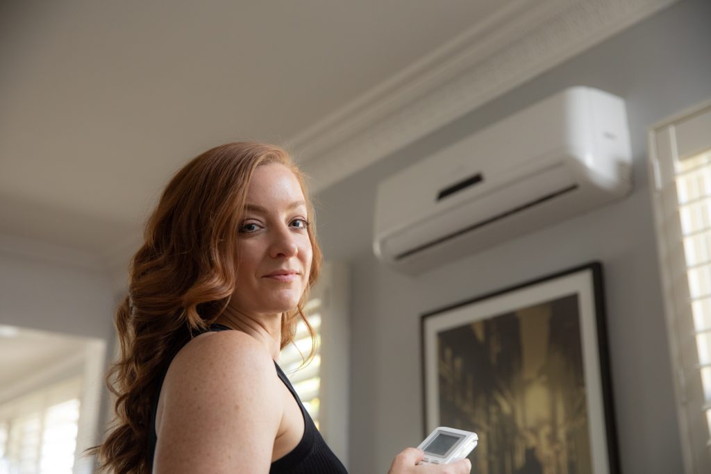 Marina Wood facing the wall, turning on an air conditioning unit, looking back smiling at the camera.