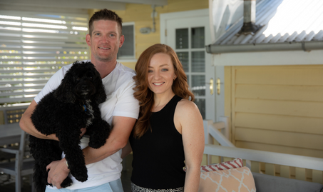 Marina and Adam Wood holding their dog, standing on their front porch smiling into the camera.