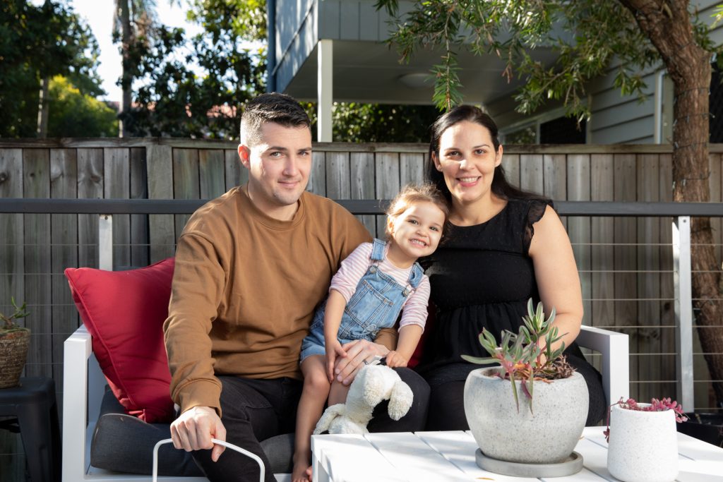 The Jackson family sitting on a couch outside, smiling into the family. A young pregnant mother, father and their toddler-aged daughter.