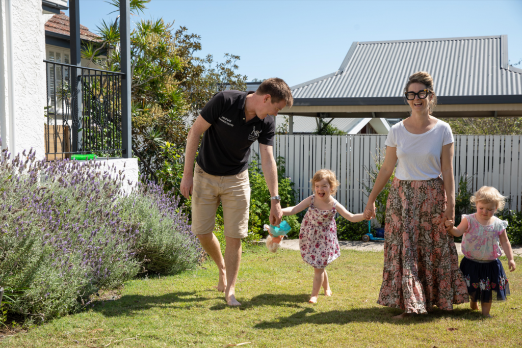 The Churton Mum and Dad holding their two young daughters hand, walking and laughing in the front yard on a sunny, blue-sky day.