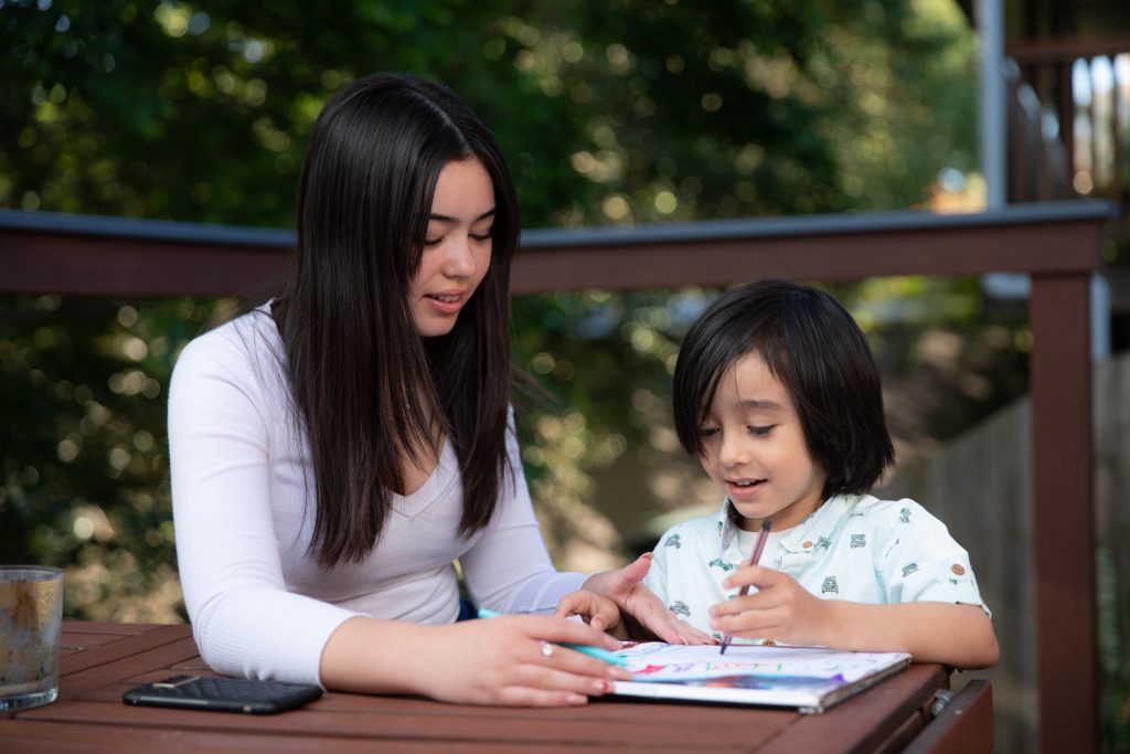 The two Hearne children sitting at a table outside with the older sister helping the younger brother colouring in.