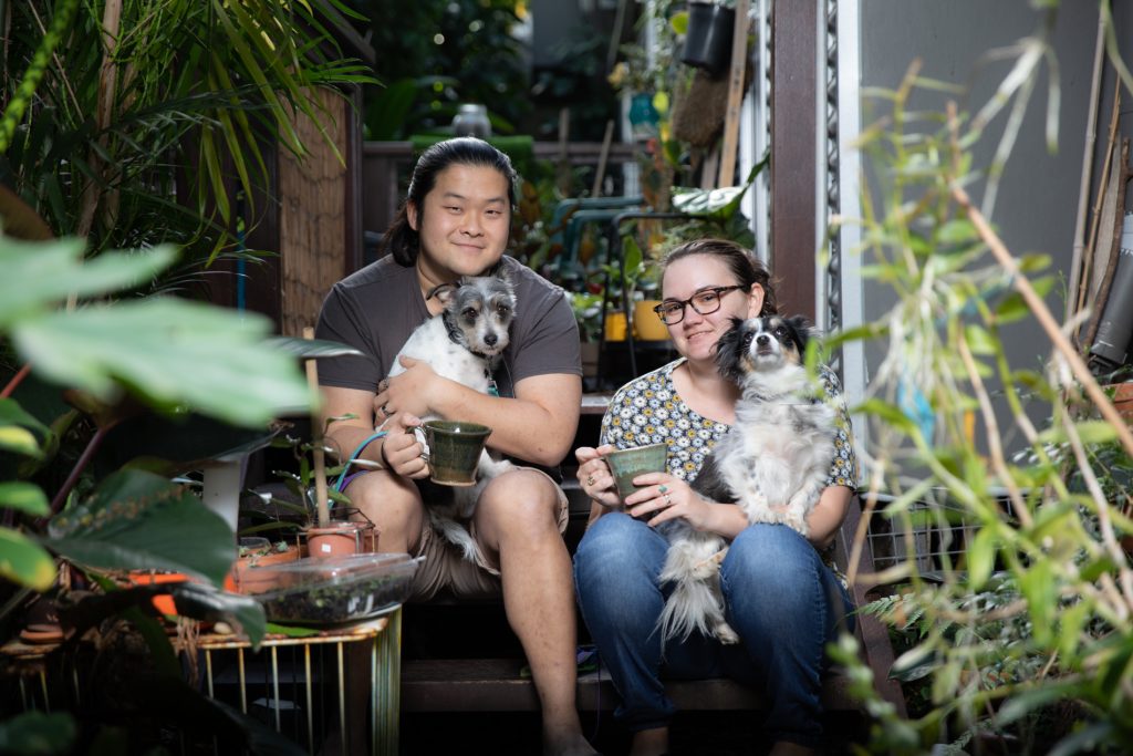Miriam Debono and Nicholas Wee sitting on steps in their courtyard with lots of surrounding green pot plants, smiling into the camera with their dogs on their laps.