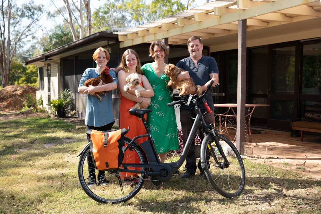 The Gittus family standing in their front yard, holding their pet dogs and chickens, with their bicycle, smiling into the camera. A Mum, Dad and a teenage daughter and son.