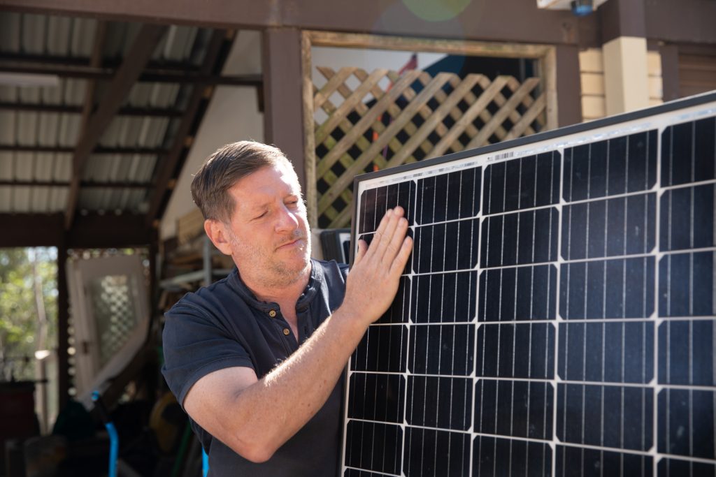 Ian Gittus, standing with his uninstalled solar panels, holding one up and inspecting it.
