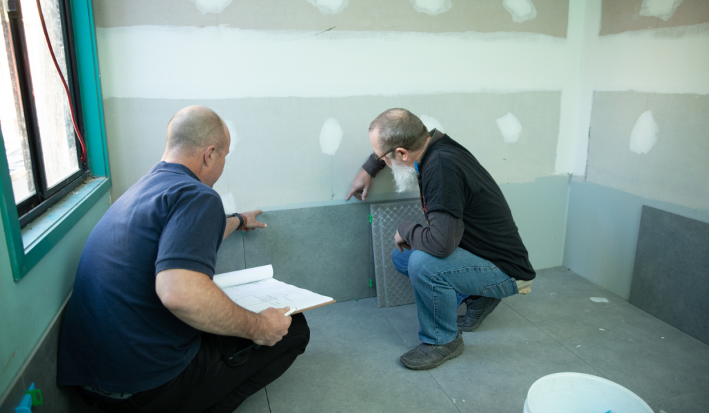 A flood resilience architect and his client with their backs turned to the camera, crouching inspecting flood resilient skirting of a house under construction.