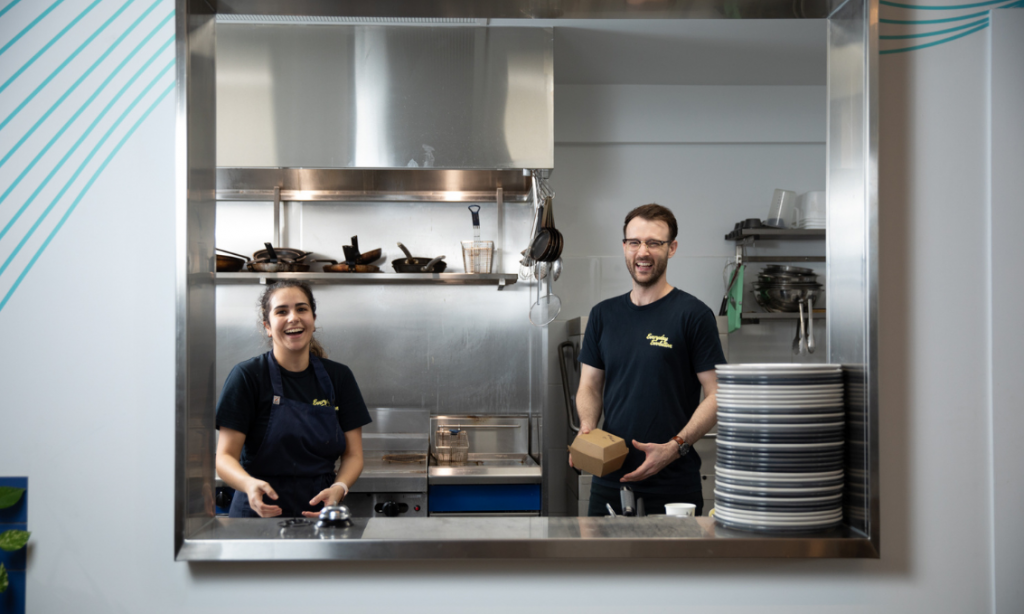 Two kitchen staff members smiling into camera, looking through window in kitchen pass