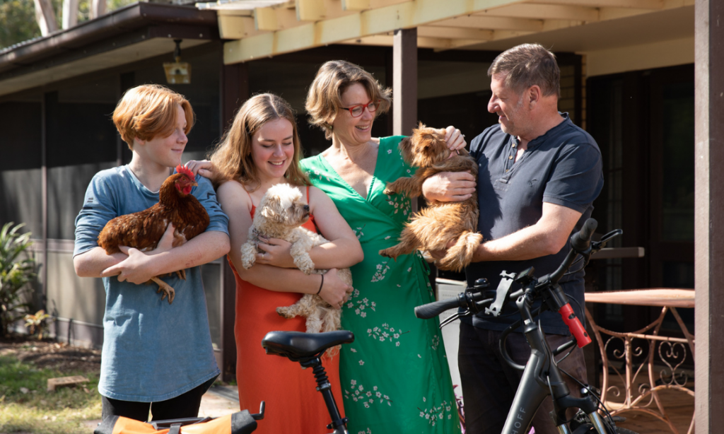 A mum, father and teenage son and daughter standing in front of their home smiling and laughing at each other while holding their family pets: a dog and two chickens.
