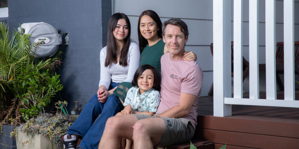 The Hearne family; husband, wife and two daughters, sitting on the stairs of their front porch, smiling into the camera.