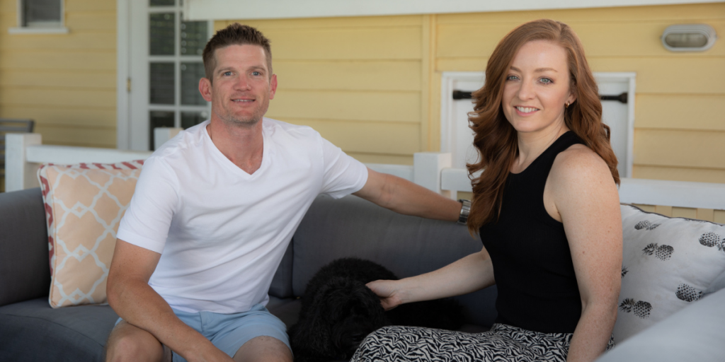 A young couple sitting on their balcony with their dog, smiling into the camera.