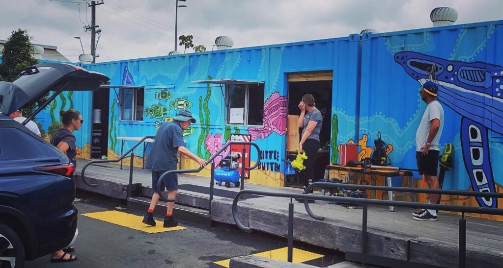 A shipping container painted in a colourful sea-themed mural, with workers and customers talking outside.
