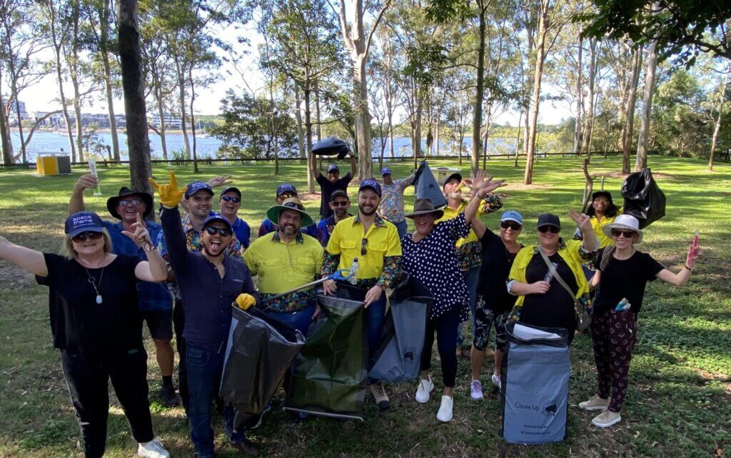 Staff members for construction company, Spaceframe, posing as a group with Clean Up Australia Day bags on the grassy bank of a beach.