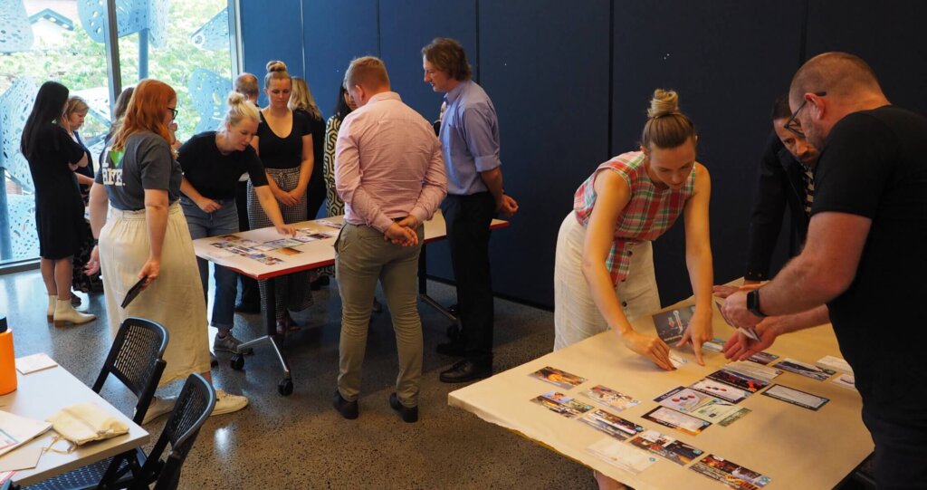 Multiple groups of 4 - 6 people standing around separate tables working together on an activity card game on the tables.